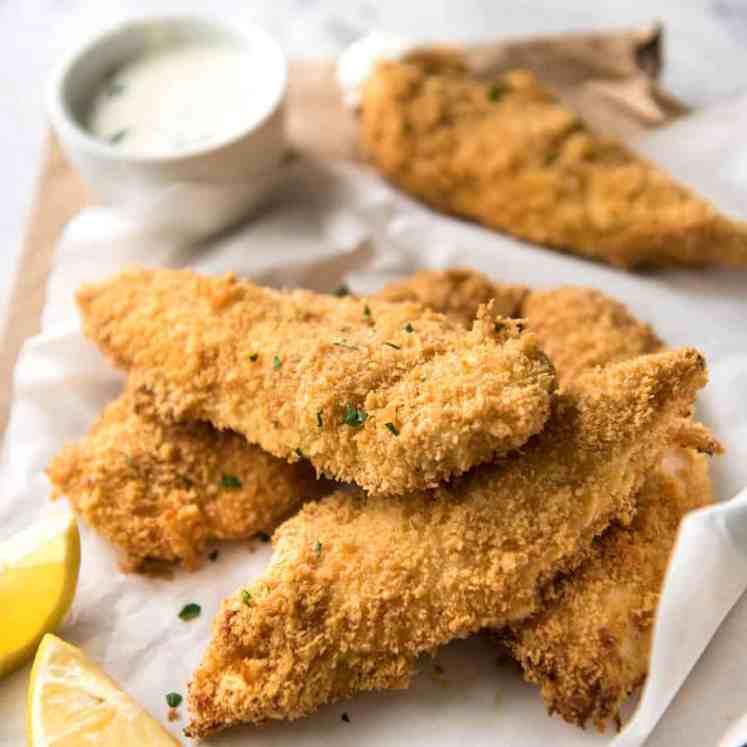 A pile of Oven Fried Chicken Tenders with lemon wedges and ranch dipping sauce in the background.